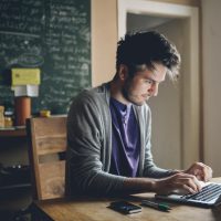 Vintage toned image of a young man working at home, freelancer, designer or writer, checking e-mails or typing on the laptop. Only natural light used, higher iso settings. Interior looks like a modern office, home or a co-working space.