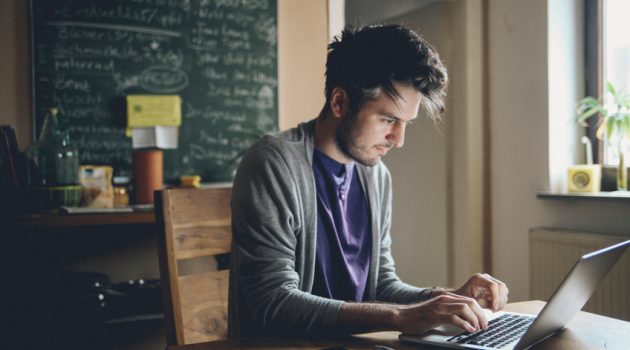 Vintage toned image of a young man working at home, freelancer, designer or writer, checking e-mails or typing on the laptop. Only natural light used, higher iso settings. Interior looks like a modern office, home or a co-working space.