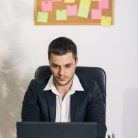 Young man, looking preoccupied and worried, sitting behind a desk in business office and working on a laptop