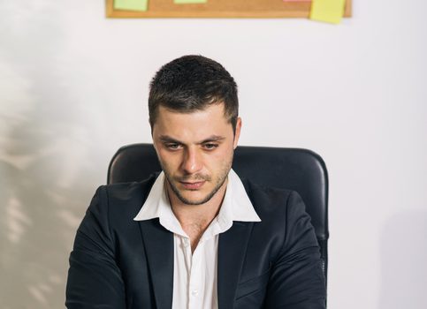 Young man, looking preoccupied and worried, sitting behind a desk in business office and working on a laptop